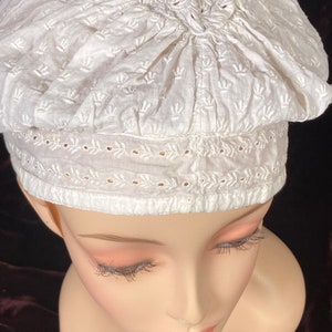 May include: White cotton bonnet with delicate embroidery. The bonnet has a gathered top and a lace trim around the bottom.