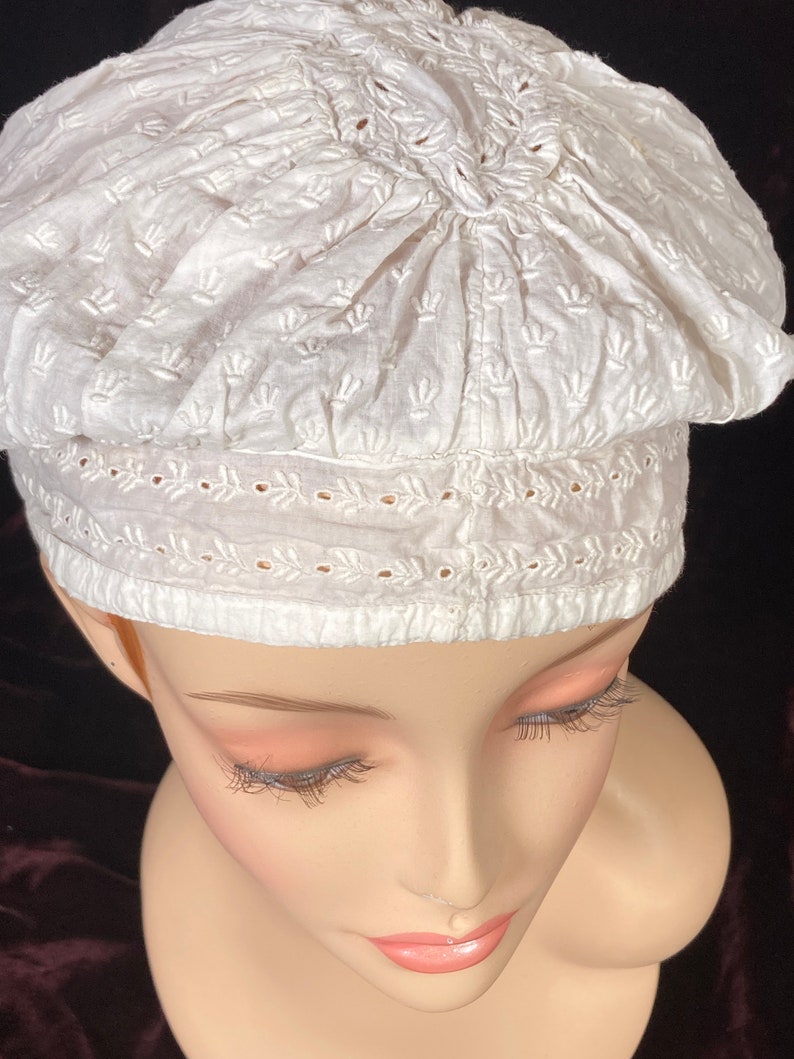 May include: White cotton bonnet with delicate embroidery. The bonnet has a gathered top and a lace trim around the bottom.
