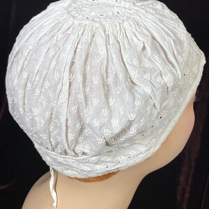 May include: White cotton bonnet with a delicate embroidered pattern. The bonnet has a gathered top and a lace trim around the edge.
