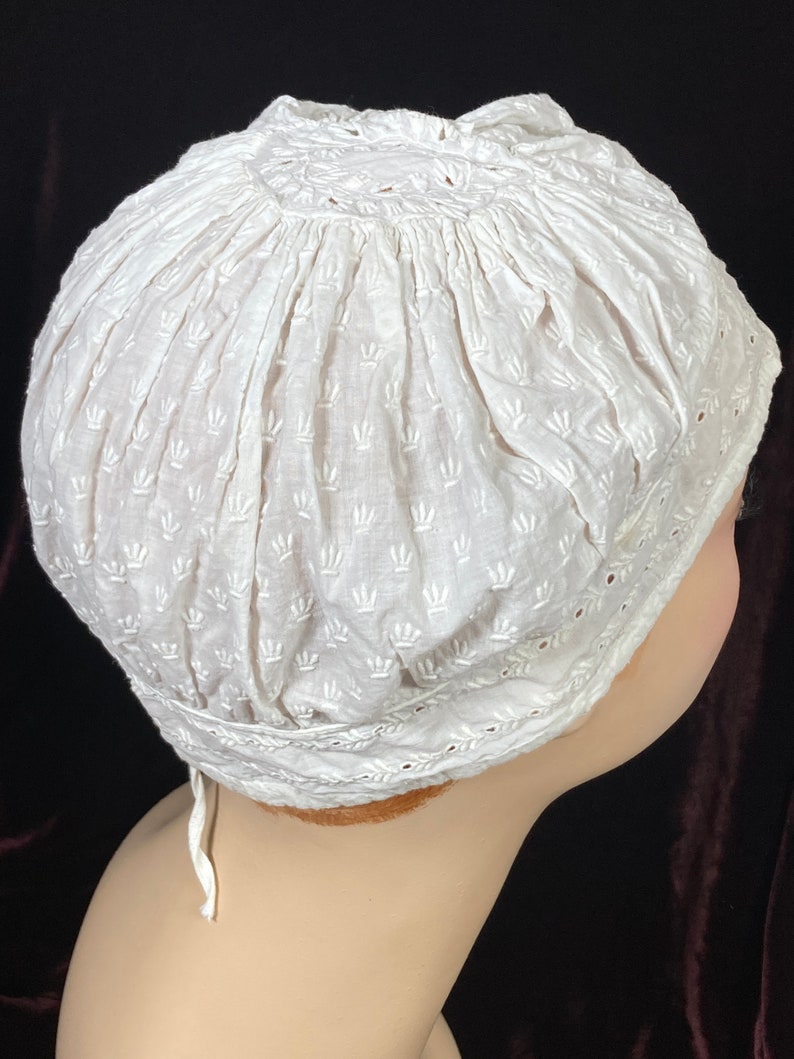 May include: White cotton bonnet with a delicate embroidered pattern. The bonnet has a gathered top and a lace trim around the edge.