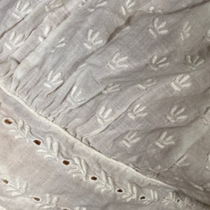 May include: White cotton fabric with a delicate embroidered pattern of small leaves. The fabric has a gathered look and a scalloped edge.