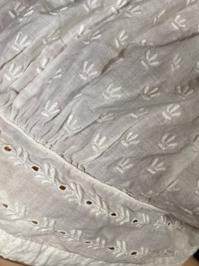 May include: White cotton fabric with a delicate embroidered pattern of small leaves. The fabric has a gathered look and a scalloped edge.