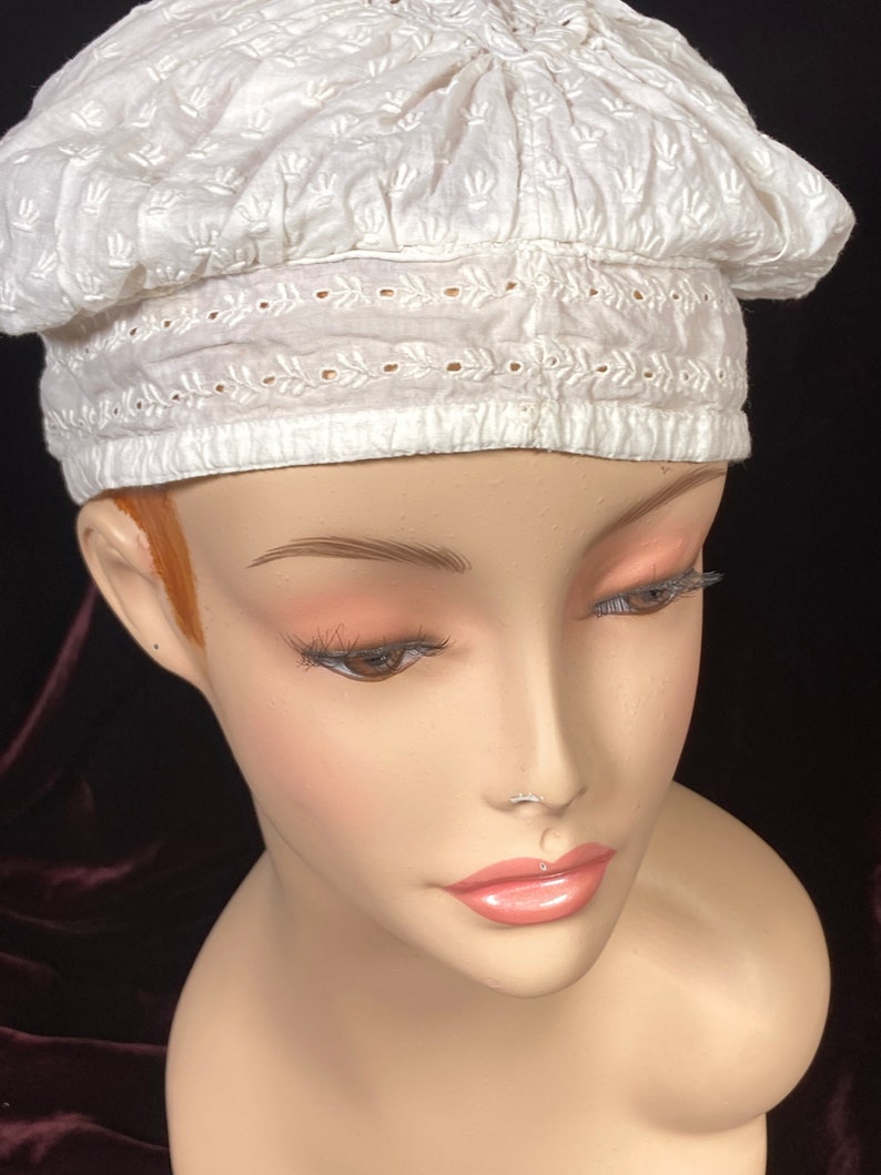 May include: White cotton beret with a delicate embroidered design. The beret is gathered at the top and has a scalloped edge.