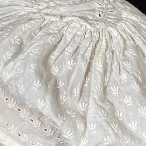 May include: White cotton cap with a gathered crown and embroidered floral design. The cap has a scalloped edge and is made of delicate fabric.