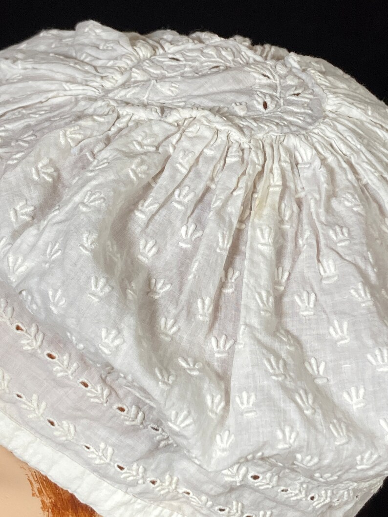 May include: White cotton cap with a gathered crown and embroidered floral design. The cap has a scalloped edge and is made of delicate fabric.