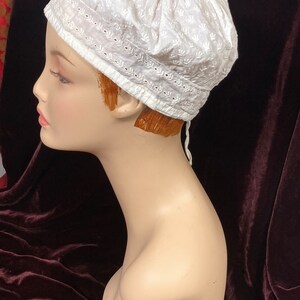 May include: White cotton cap with a delicate embroidered design. The cap has a rounded shape and a small tie at the back.