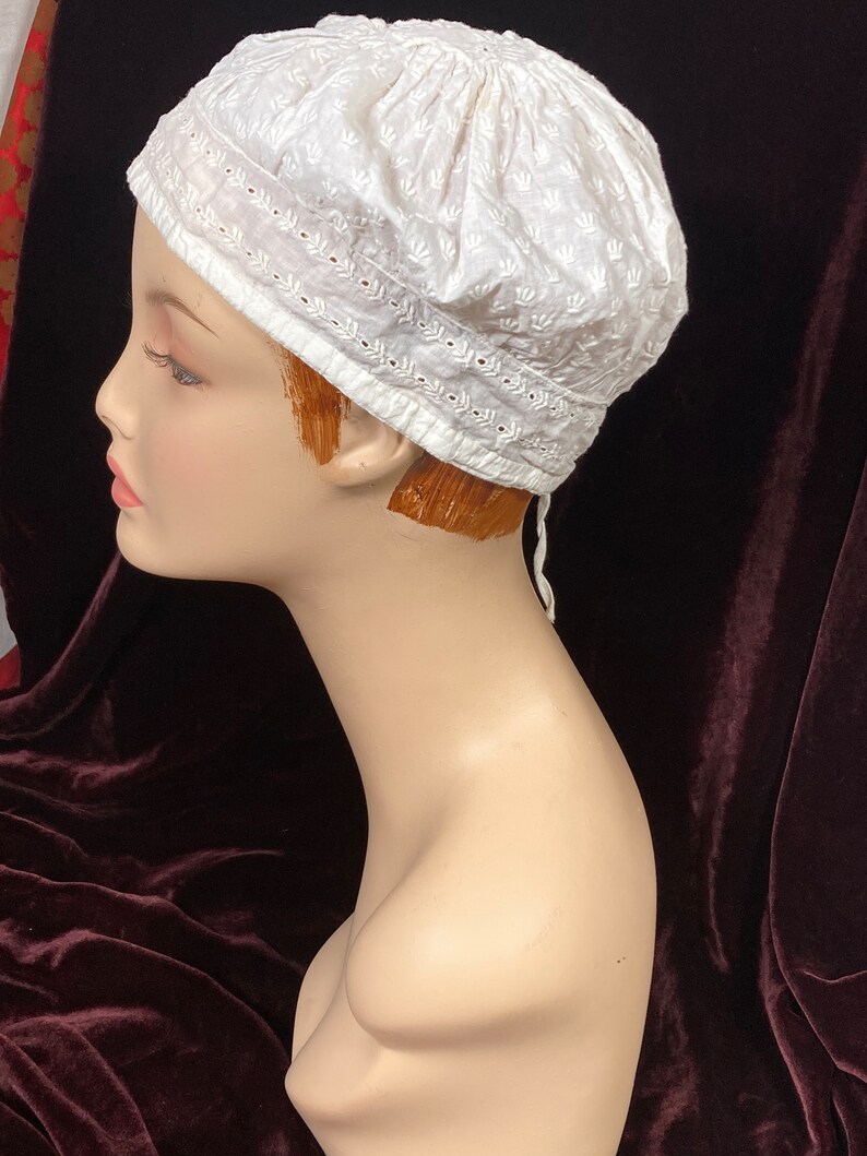 May include: White cotton cap with a delicate embroidered design. The cap has a rounded shape and a small tie at the back.