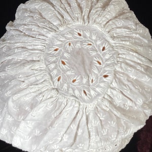 May include: White cotton bonnet with a delicate floral embroidery pattern. The bonnet has a gathered ruffle around the edge and a circular design in the center.