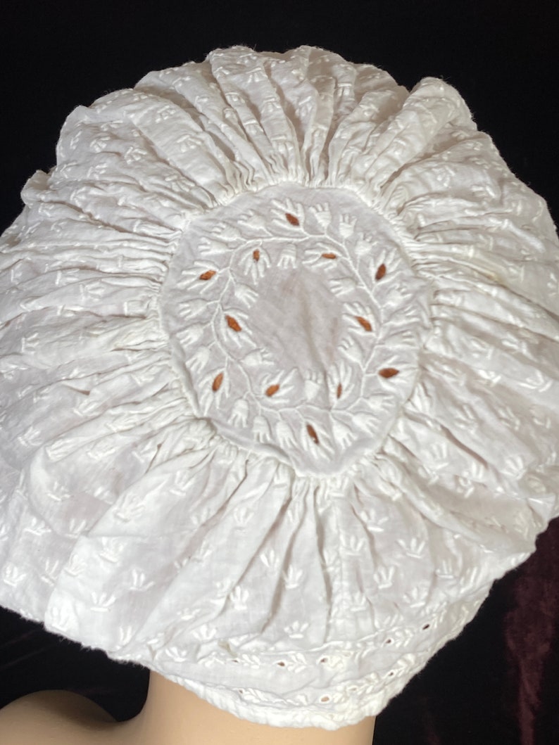 May include: White cotton bonnet with a delicate floral embroidery pattern. The bonnet has a gathered ruffle around the edge and a circular design in the center.