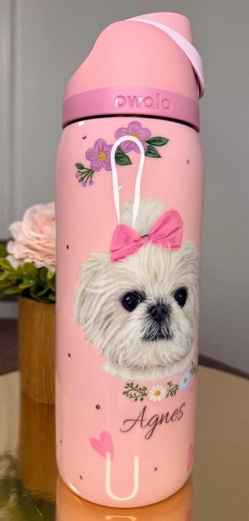Op de afbeelding: Roze waterfles met een illustratie van een witte hond met een roze strik. De fles heeft het woord "owala" op het deksel en de naam "Agnes" op de bodem. Bloemen- en hartaccenten versieren de fles.