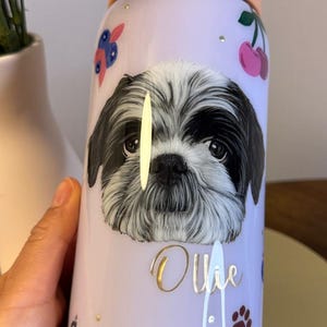 Op de afbeelding: Een lichtpaarse Owala waterfles met een zwart-witte Shih Tzu hondenillustratie. De fles heeft gouden letters met de tekst "Ollie", samen met kersen- en pootafdrukontwerpen. De fles heeft een perzik-kleurige handgreep.