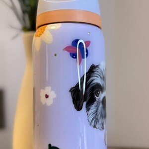 May include: A lavender Owala water bottle with a dog illustration, flowers, and cherries. The bottle has a light blue lid and a peach-colored handle. The Owala logo is printed on the side.