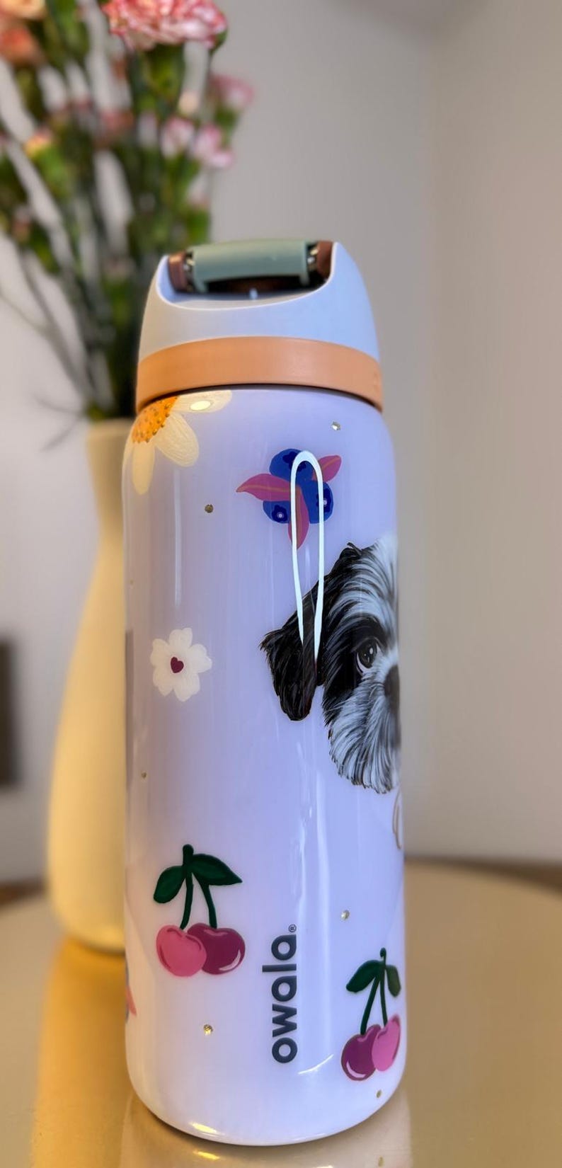 May include: A lavender Owala water bottle with a dog illustration, flowers, and cherries. The bottle has a light blue lid and a peach-colored handle. The Owala logo is printed on the side.