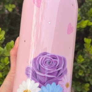 May include: A pink Owala water bottle with a floral design. The bottle features a large purple rose, white and blue daisies, and pink heart accents. The bottle is cylindrical and has a pink lid.