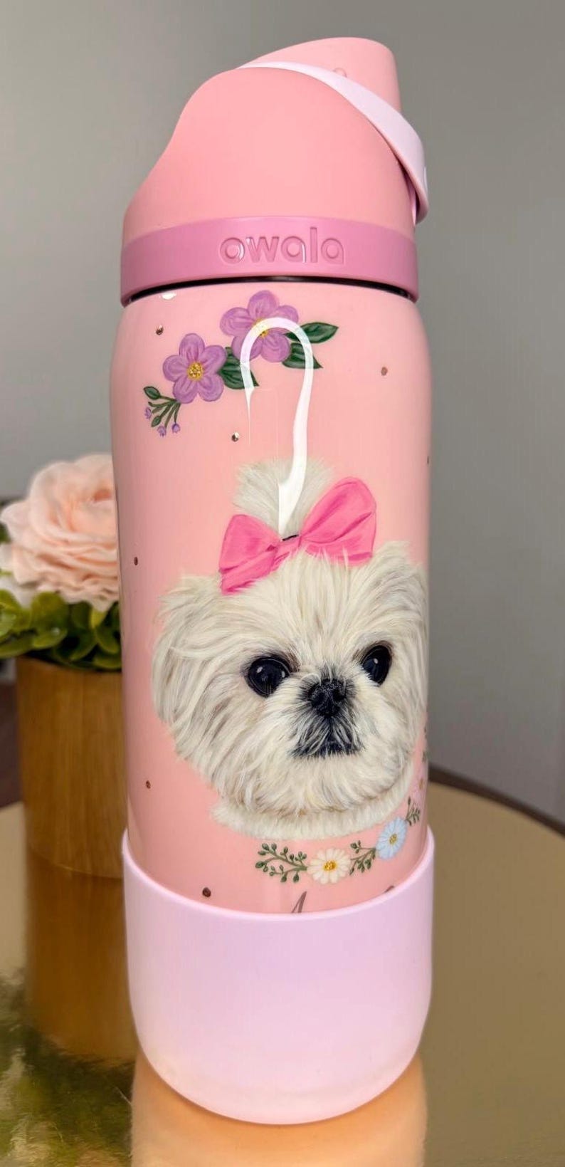 Op de afbeelding: Roze ge&iuml;soleerde waterfles met een illustratie van een witte hond met een roze strik. De fles heeft een roze deksel en basis, met de merknaam "owala" erop gedrukt. Bloemen- en stipaccenten versieren de fles.