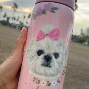 Op de afbeelding: Roze Owala waterfles met een witte hond met een roze strik, bloemaccenten en de naam "Agnes". De fles heeft een roze deksel en handvat. De fles is ontworpen om drankjes koud te houden.