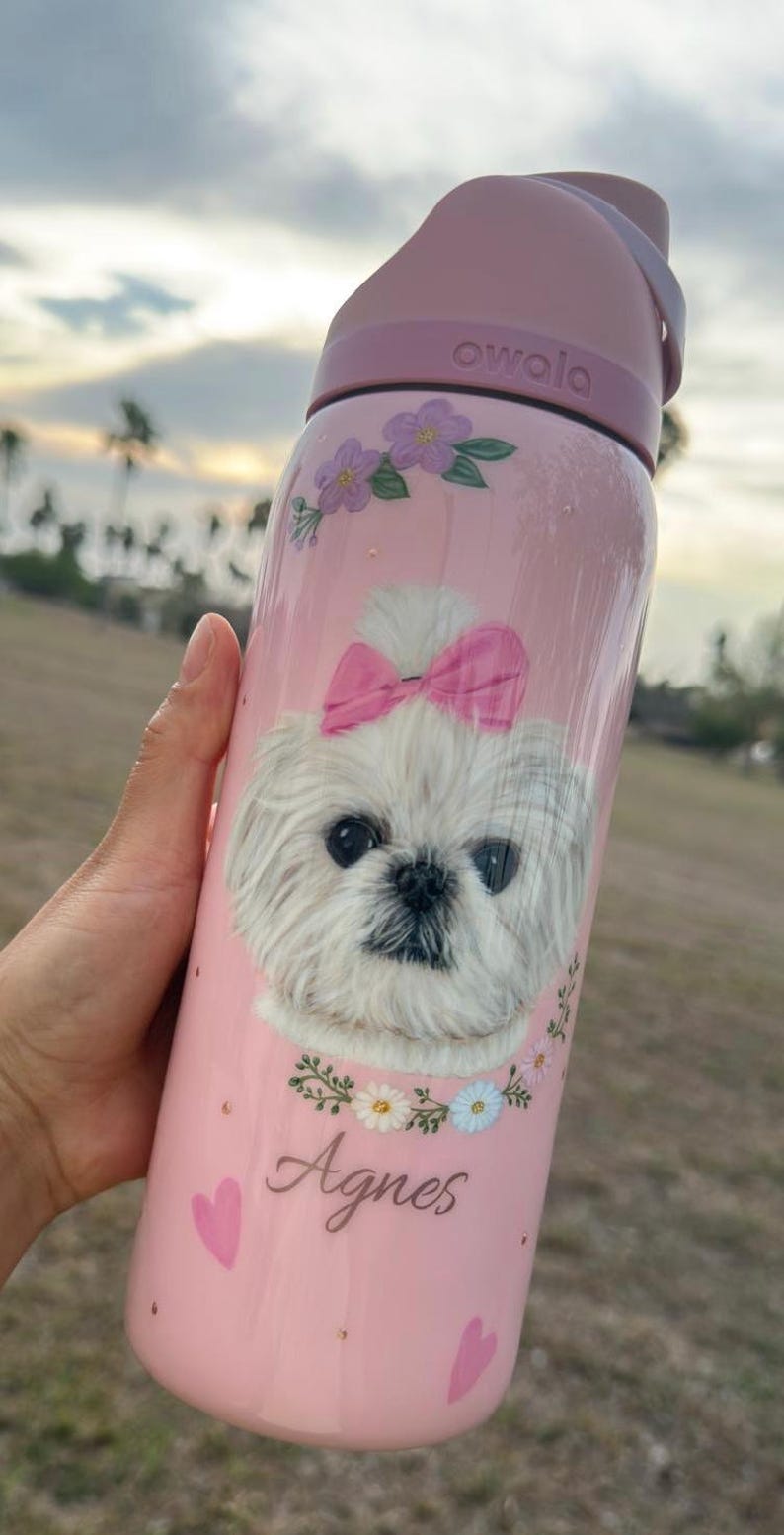 Op de afbeelding: Roze Owala waterfles met een witte hond met een roze strik, bloemaccenten en de naam "Agnes". De fles heeft een roze deksel en handvat. De fles is ontworpen om drankjes koud te houden.
