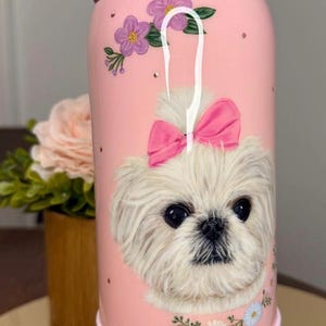 May include: Pink Owala water bottle featuring a white dog with a pink bow, surrounded by purple flowers and gold dots. The bottle has a pink handle and base, with the brand name "owala" on the lid.