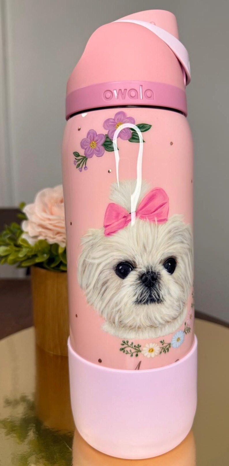 May include: Pink Owala water bottle featuring a white dog with a pink bow, surrounded by purple flowers and gold dots. The bottle has a pink handle and base, with the brand name "owala" on the lid.