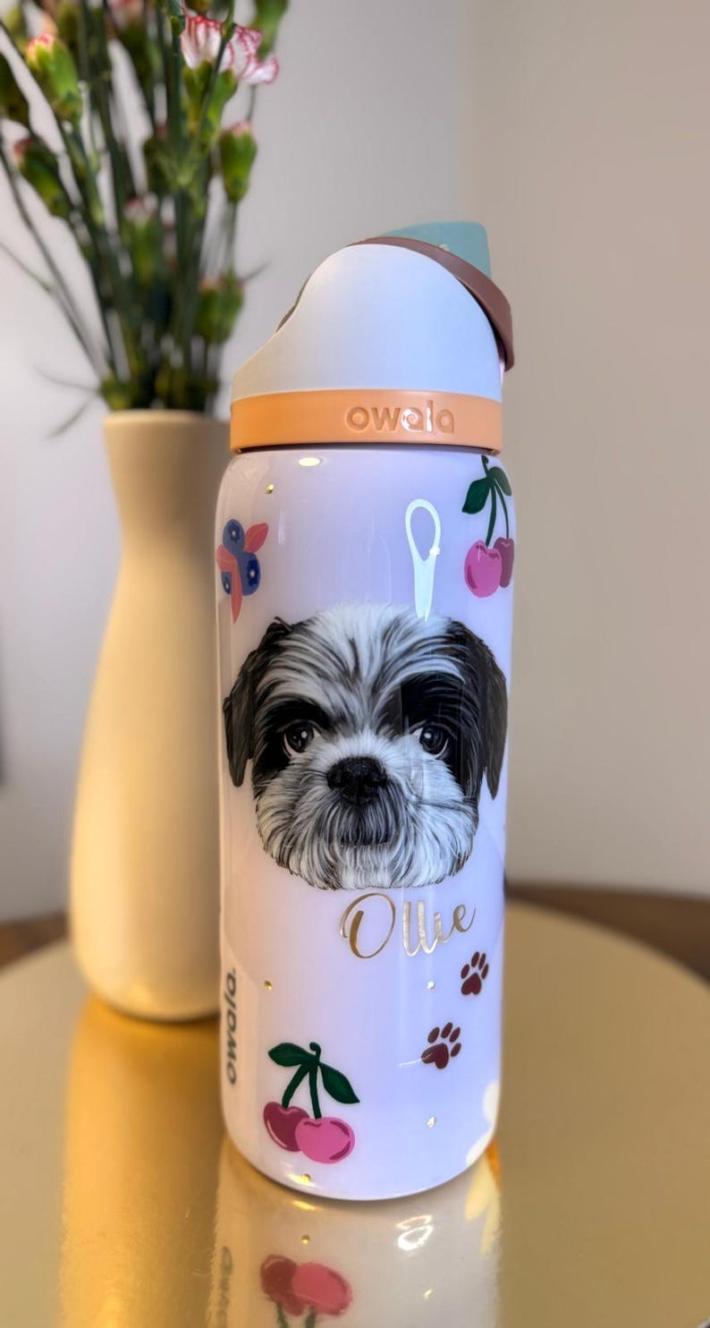 May include: A light purple Owala water bottle featuring a Shih Tzu dog portrait, the name "Ollie", cherries, paw prints, and floral accents. The bottle has a white and orange lid.