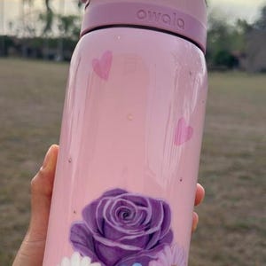 Op de afbeelding: Roze Owala waterfles met een paarse roos en madeliefjes bloemenontwerp. De fles heeft een roze deksel en het woord "owala" staat erop gedrukt. Twee roze harten zijn ook op de fles geschilderd.