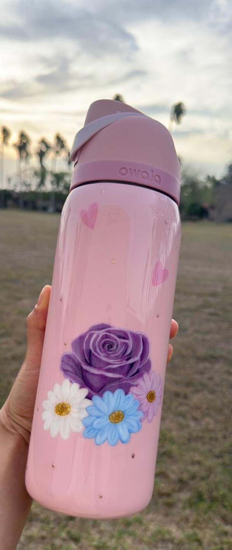 Op de afbeelding: Roze Owala waterfles met een paarse roos en madeliefjes bloemenontwerp. De fles heeft een roze deksel en het woord "owala" staat erop gedrukt. Twee roze harten zijn ook op de fles geschilderd.