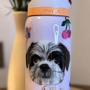 Op de afbeelding: Een lichtpaarse Owala waterfles met een Shih Tzu hondenillustratie, de naam "Ollie" en kersen- en pootafdrukontwerpen. De fles heeft een witte en oranje dop met een bruin handvat.