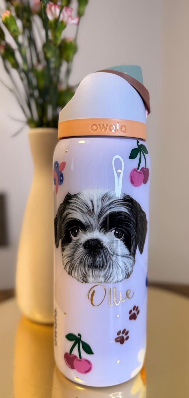 Op de afbeelding: Een lichtpaarse Owala waterfles met een Shih Tzu hondenillustratie, de naam "Ollie" en kersen- en pootafdrukontwerpen. De fles heeft een witte en oranje dop met een bruin handvat.