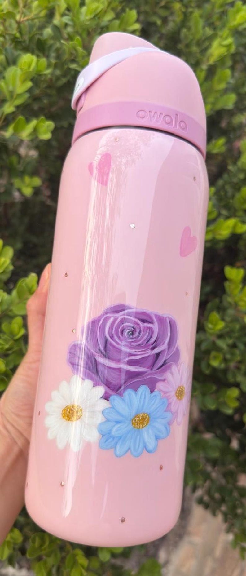 Op de afbeelding: Een roze Owala waterfles met een bloemenontwerp. De fles heeft een grote paarse roos, witte en blauwe madeliefjes en roze hartaccenten. De fles is cilindrisch en heeft een roze deksel.