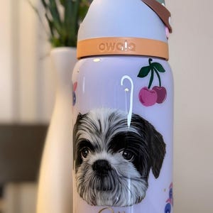 May include: A lilac-colored Owala water bottle featuring a Shih Tzu dog portrait, cherries, and paw prints. The bottle has a light blue lid and a brown handle. The word "Ollie" is written in gold script.