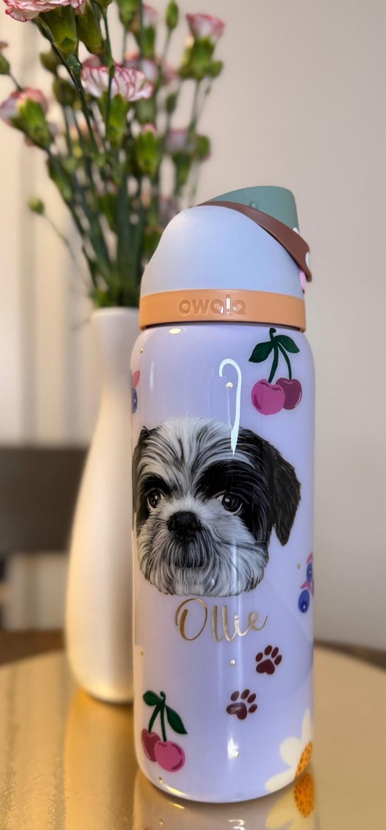 May include: A lilac-colored Owala water bottle featuring a Shih Tzu dog portrait, cherries, and paw prints. The bottle has a light blue lid and a brown handle. The word "Ollie" is written in gold script.