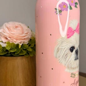 Op de afbeelding: Roze waterfles met een hondenillustratie. De fles heeft een witte hond met een roze strik, bloemen en de naam "Agnes". De fles heeft een roze handvat en deksel en is versierd met gouden spikkels.