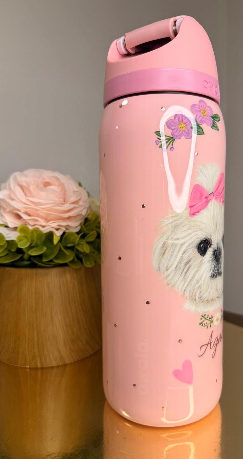 Op de afbeelding: Roze waterfles met een hondenillustratie. De fles heeft een witte hond met een roze strik, bloemen en de naam "Agnes". De fles heeft een roze handvat en deksel en is versierd met gouden spikkels.