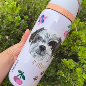 Op de afbeelding: Witte Owala waterfles met een Shih Tzu hondenportret, de naam "Ollie" in gouden letters en roze kersen. De fles heeft een perzikkleurige handgreep en deksel, met de merknaam "owala" aan de zijkant.