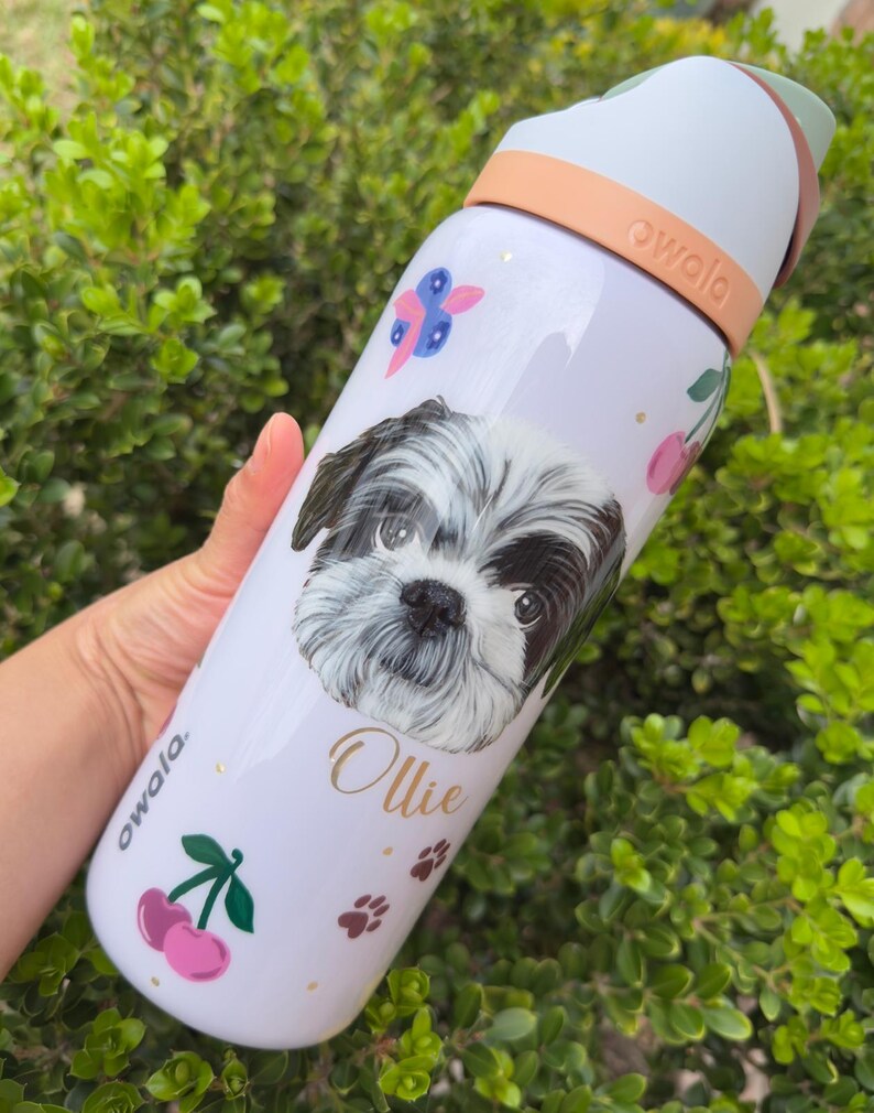 Op de afbeelding: Witte Owala waterfles met een Shih Tzu hondenportret, de naam "Ollie" in gouden letters en roze kersen. De fles heeft een perzikkleurige handgreep en deksel, met de merknaam "owala" aan de zijkant.
