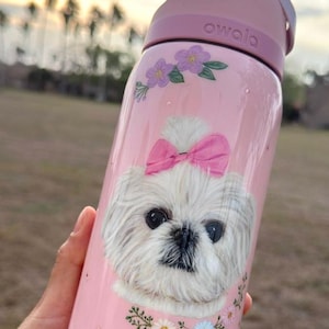 May include: Pink Owala water bottle featuring a white dog illustration with a pink bow, floral accents, and the name "Agnes." The bottle has a pink lid and handle.