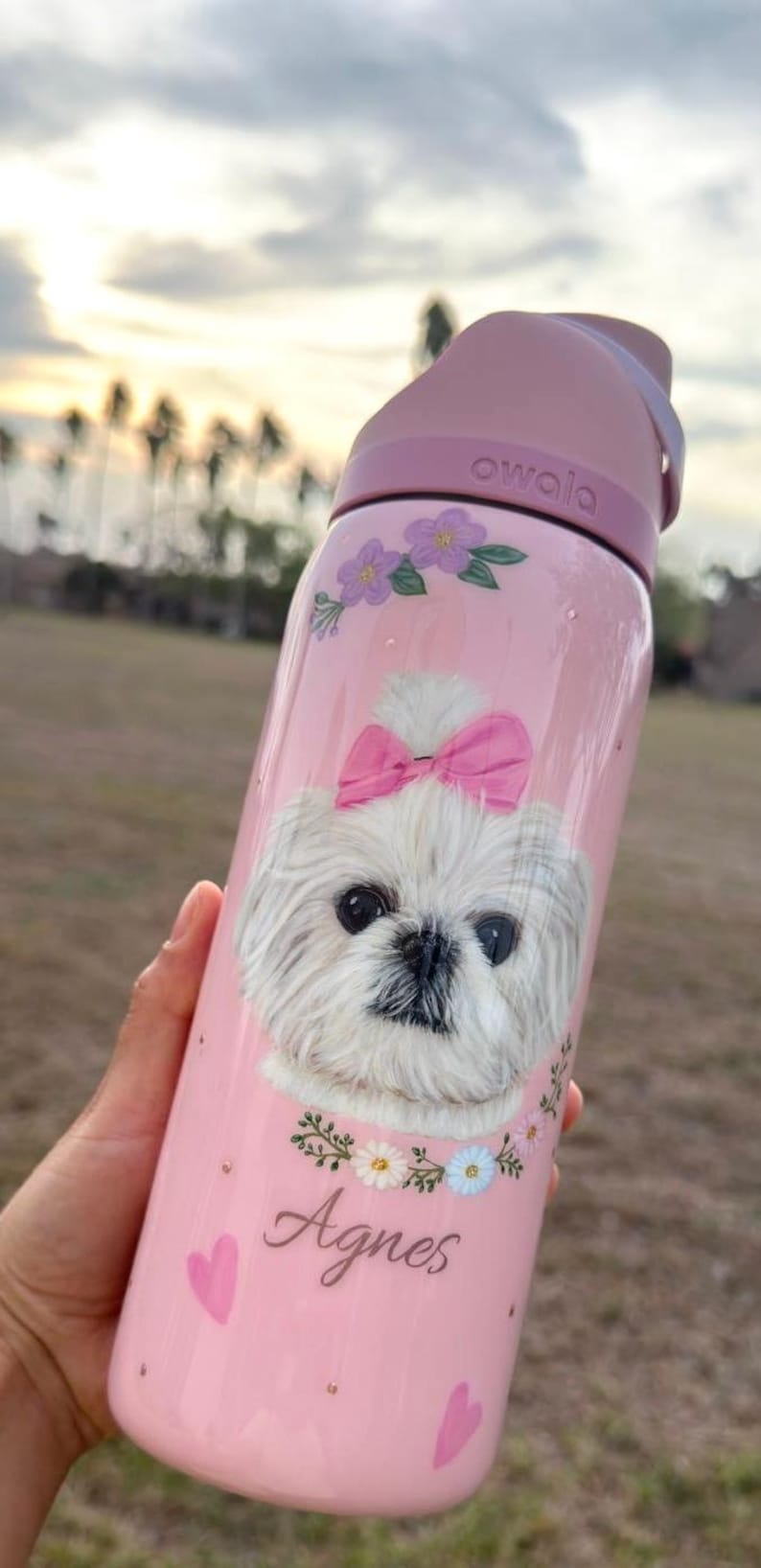 May include: Pink Owala water bottle featuring a white dog illustration with a pink bow, floral accents, and the name "Agnes." The bottle has a pink lid and handle.