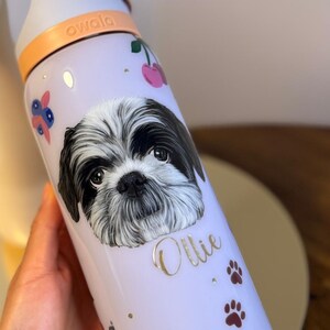 Op de afbeelding: Een lichtpaarse Owala waterfles met een Shih Tzu hondenillustratie en de naam "Ollie". De fles heeft een witte dop en een oranje handvat. Versierd met kersen, pootafdrukken en bloemaccenten.