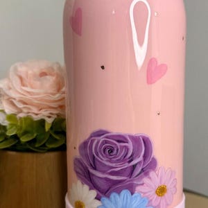 May include: Pink Owala water bottle with a floral design. The bottle features a large purple rose and several smaller daisies in white, blue, and pink. The bottle also has pink hearts and gold flecks.