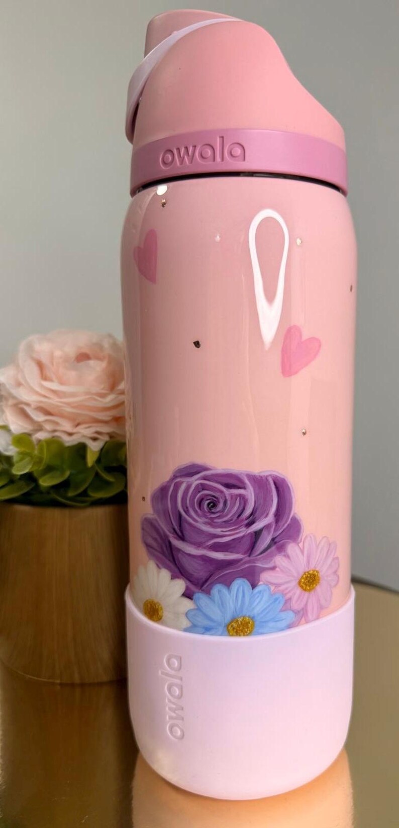 May include: Pink Owala water bottle with a floral design. The bottle features a large purple rose and several smaller daisies in white, blue, and pink. The bottle also has pink hearts and gold flecks.