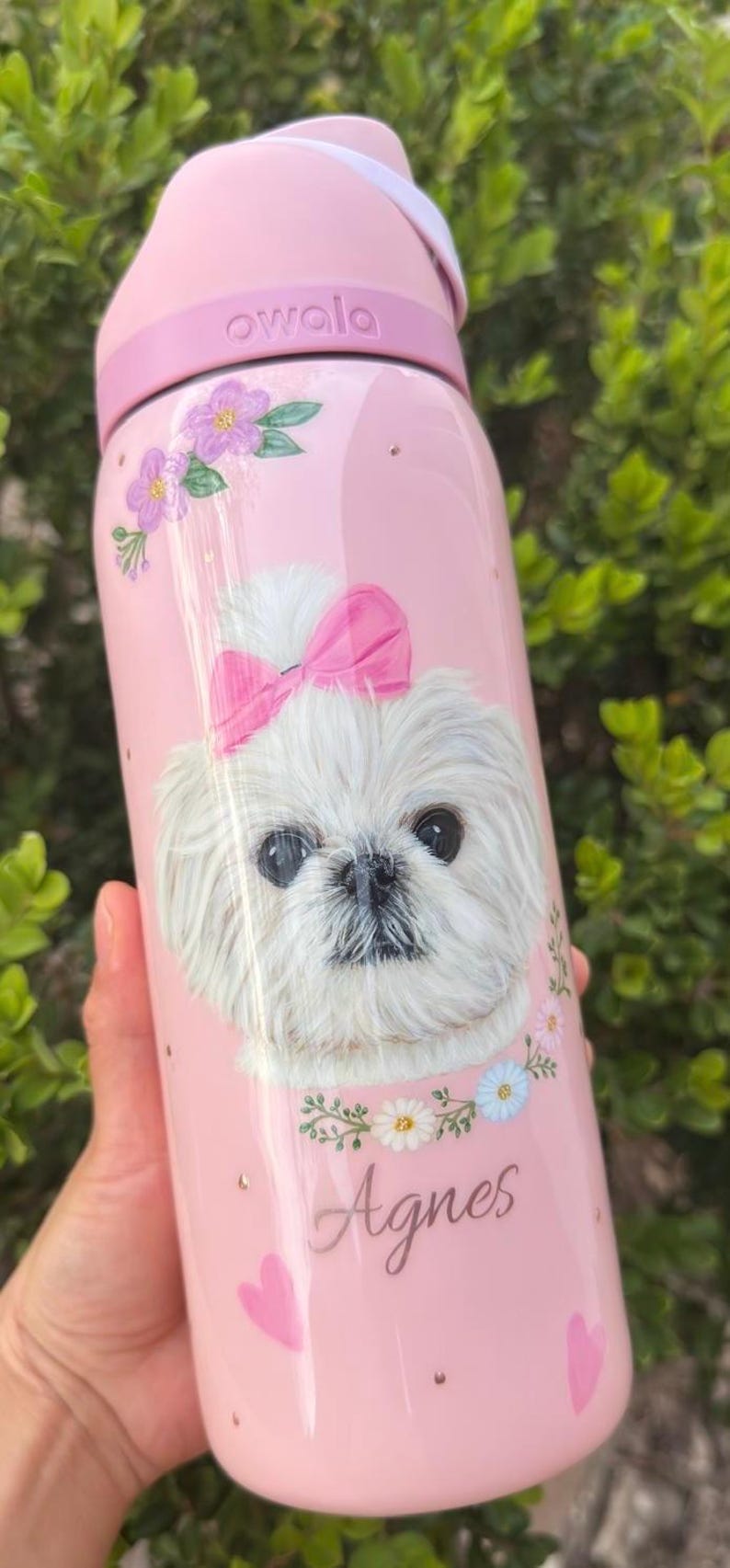 Op de afbeelding: Roze Owala waterfles met een illustratie van een witte hond met een roze strik, bloemen en de naam "Agnes". De fles heeft een roze deksel en handvat en is versierd met kleine gouden stippen.