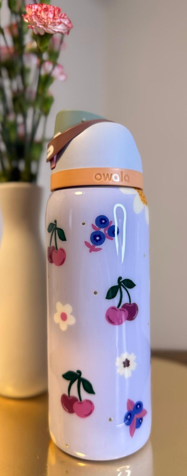 May include: A light purple Owala water bottle with a white lid and handle. The bottle is decorated with cherries, blueberries, and white flowers. The word "owala" is printed on the bottle.