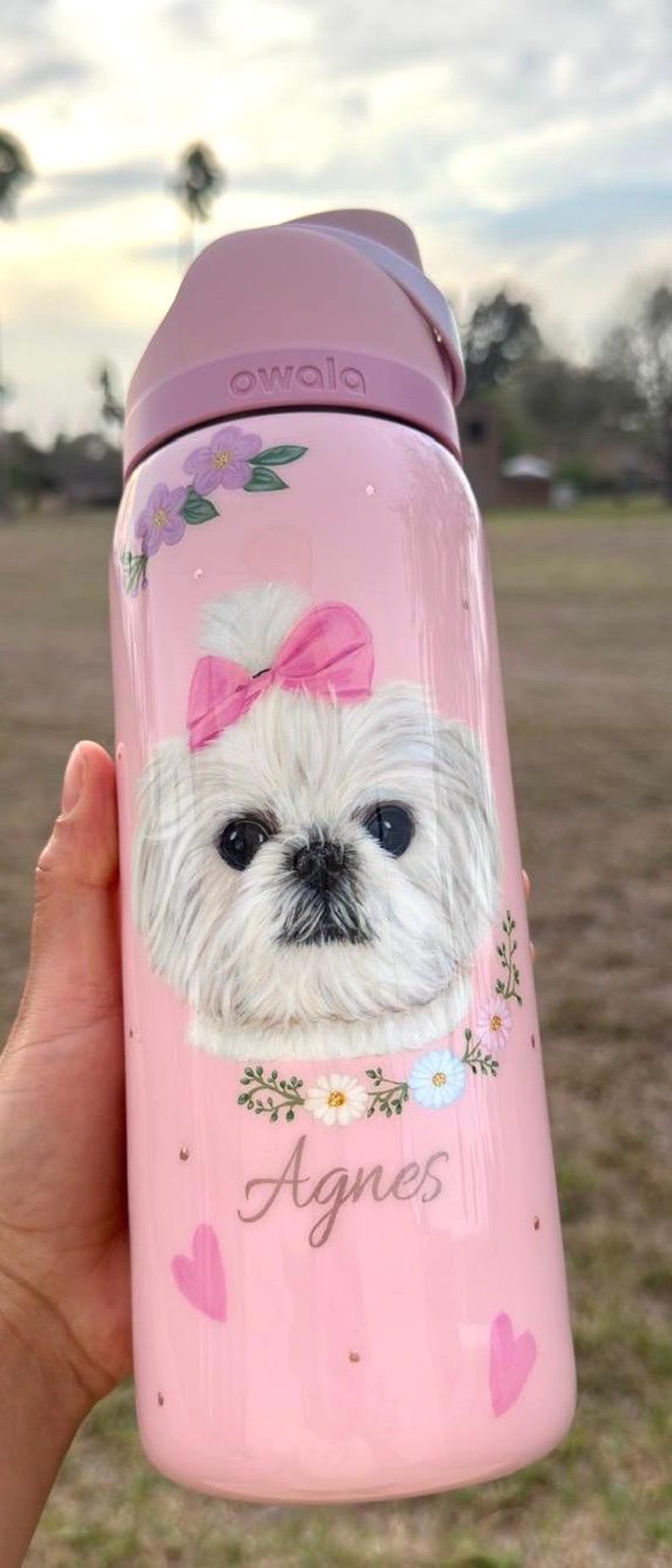 Op de afbeelding: Roze Owala waterfles met een illustratie van een witte hond met een roze strik. De fles is versierd met bloemaccenten en de naam "Agnes".