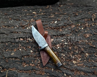 May include: A Damascus steel knife with a wooden handle and a leather sheath. The blade has a unique pattern, and the handle is a light brown color. The knife is resting on a dark brown, textured surface.