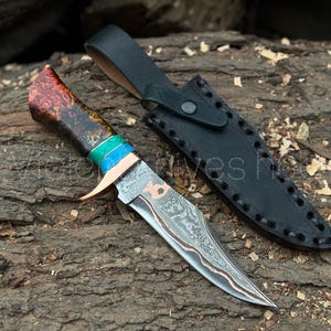 Könnte beinhalten: Ein handgefertigtes Messer mit gemusterter Klinge und einem Griff mit roten, grünen und blauen Akzenten. Das Messer wird von einer schwarzen Lederscheide begleitet, die auf einer strukturierten Holzoberfläche liegt.