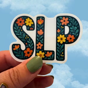 May include: A white sticker with the letters "SLP" in a bold, floral design. The letters are outlined in dark teal and filled with orange and yellow flowers and green leaves. The sticker is held against a blue sky background.