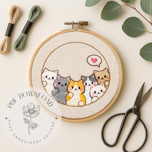 May include: An embroidery hoop featuring a hand-stitched design of seven cartoon cats in various colors. The cats are arranged in a semi-circle, with a heart-shaped speech bubble above them. Two spools of thread and a pair of scissors are also visible.