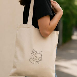 以下が含まれることがあります： ベージュ色のキャンバス地のトートバッグ。頭巾をかぶった猫の線画が描かれています。猫は不機嫌そうな表情をしています。バッグには長いストラップが付いており、肩にかけています。
