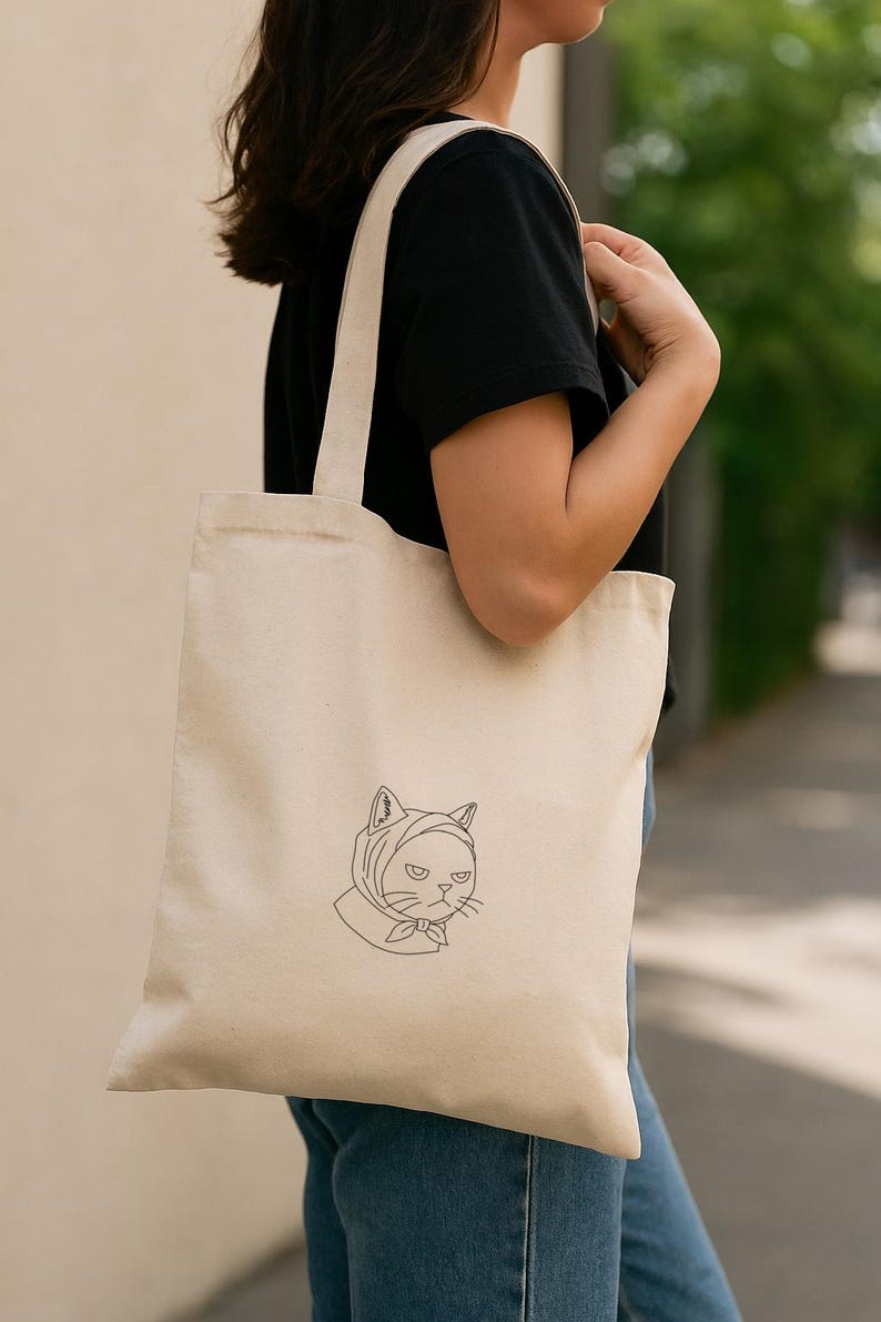 以下が含まれることがあります： ベージュ色のキャンバス地のトートバッグ。頭巾をかぶった猫の線画が描かれています。猫は不機嫌そうな表情をしています。バッグには長いストラップが付いており、肩にかけています。