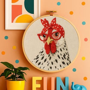 May include: A circular embroidery hoop featuring a stitched image of a white chicken with black spots, wearing red glasses and a red polka-dot headscarf. The chicken has a yellow beak and red wattle, set against a light beige fabric background.